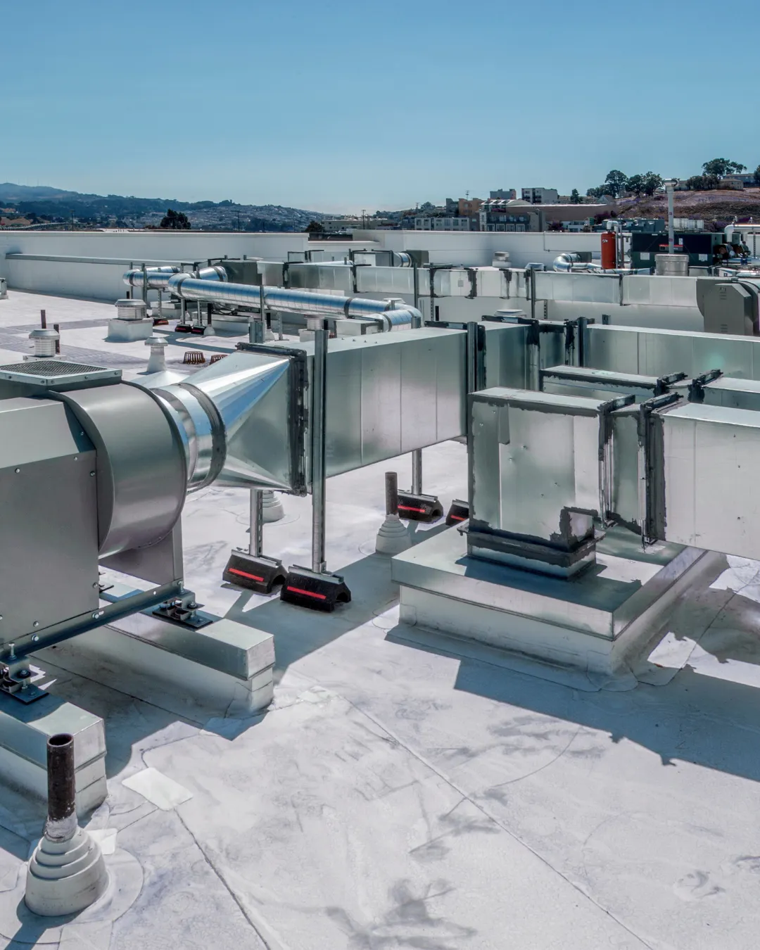 Industrial rooftop HVAC system with metal ductwork, equipment units, and mechanical installations overlooking a bay with hills and residential areas in the background