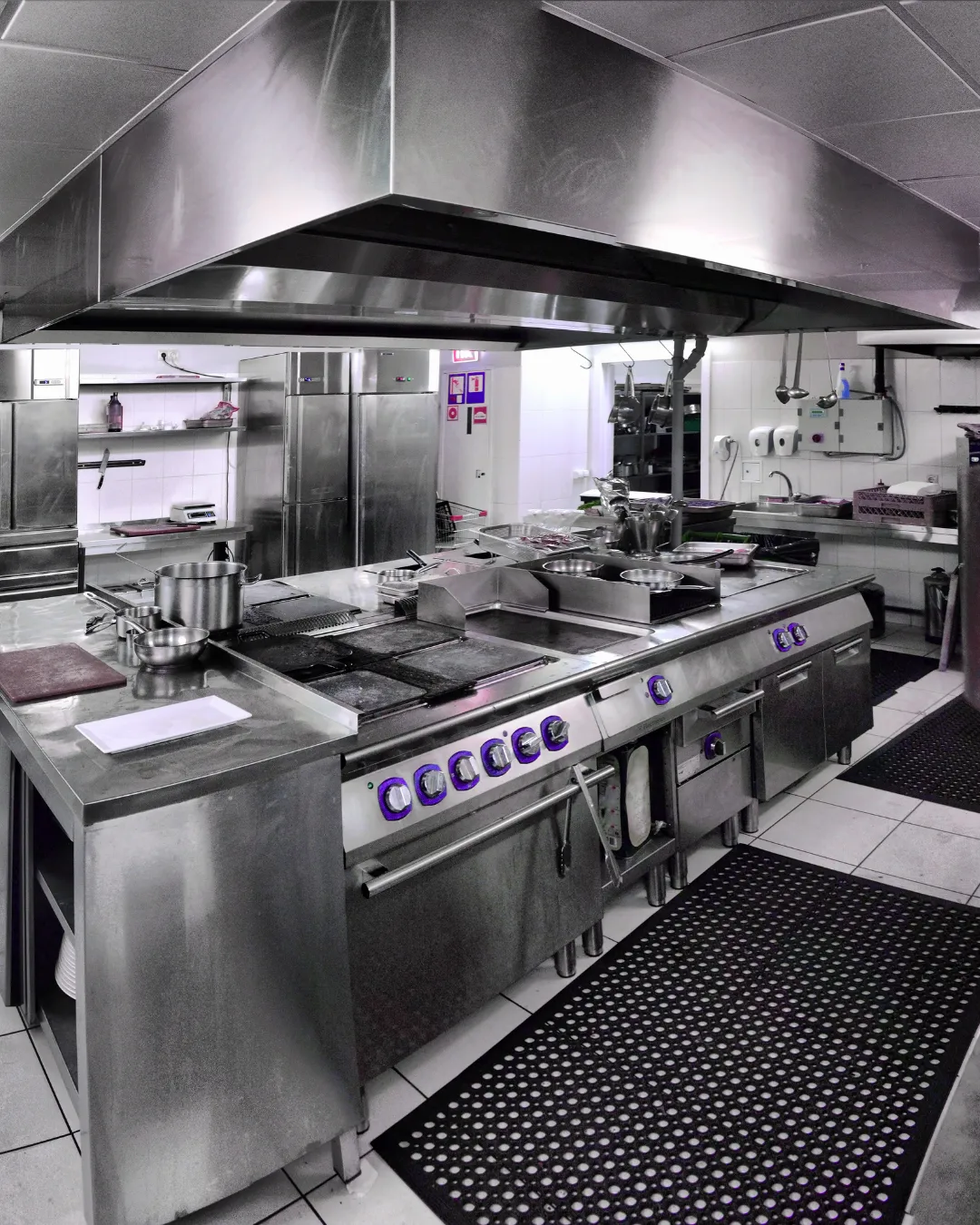 Professional commercial kitchen interior with stainless steel industrial cooking equipment, overhead ventilation hood, and black perforated rubber flooring