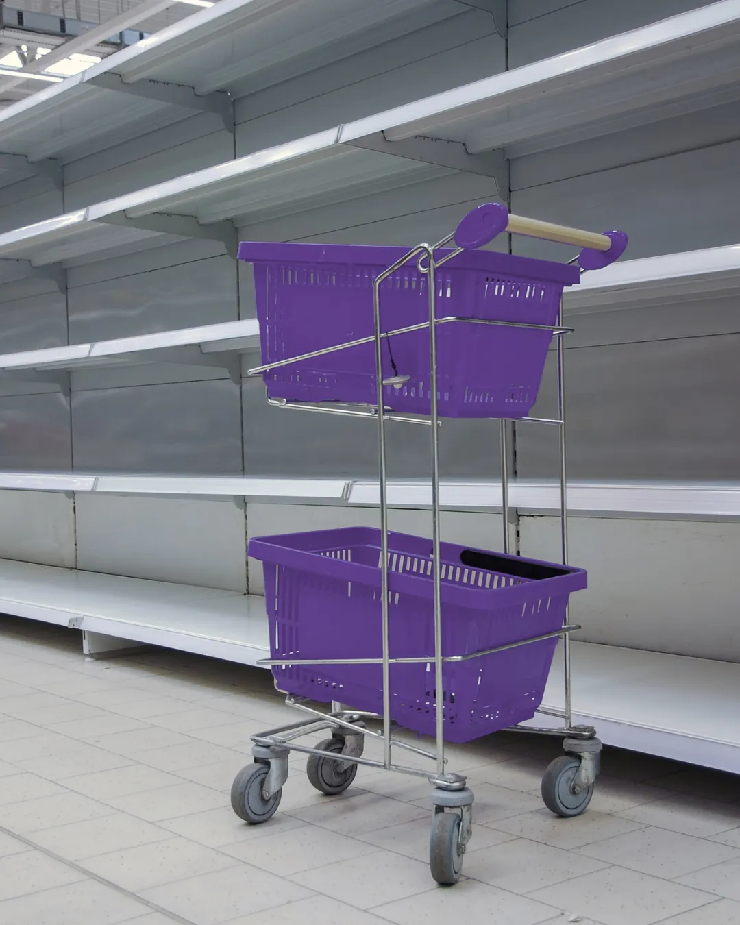 Purple shopping baskets stacked on a metal rolling cart in an empty retail store with fluorescent ceiling lights and white shelving units in the background