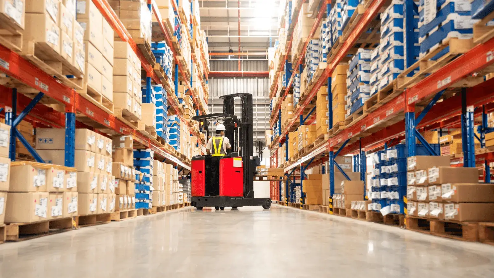 A warehouse aisle with high pallet racks filled with boxes and a person operating a red forklift.