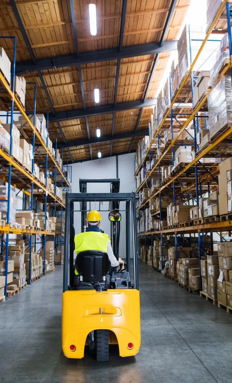 Warehouse scene with a worker driving a forklift between tall pallet racks filled with boxes.