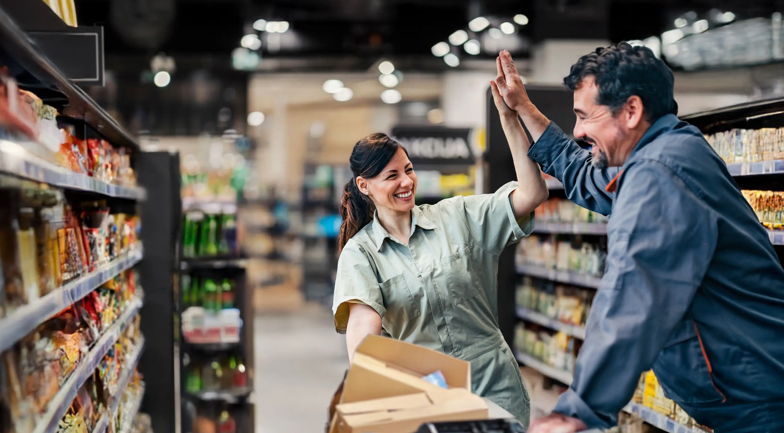 Two smiling supermarket employees high-fiving in an aisle surrounded by shelves stocked with products.