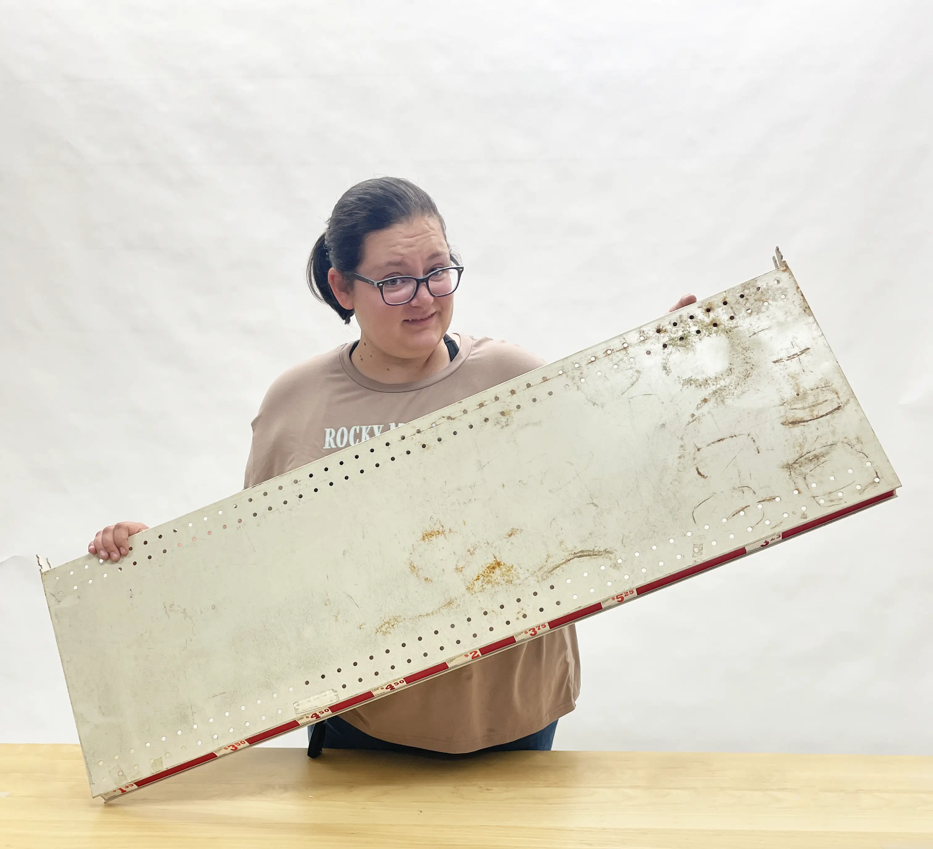 Person holding a large, worn piece of retail shelving with holes, standing in front of a plain background.