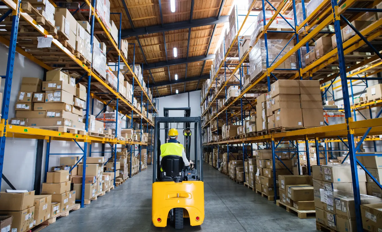 A person operating a forklift in a large warehouse with shelves stacked with boxes.