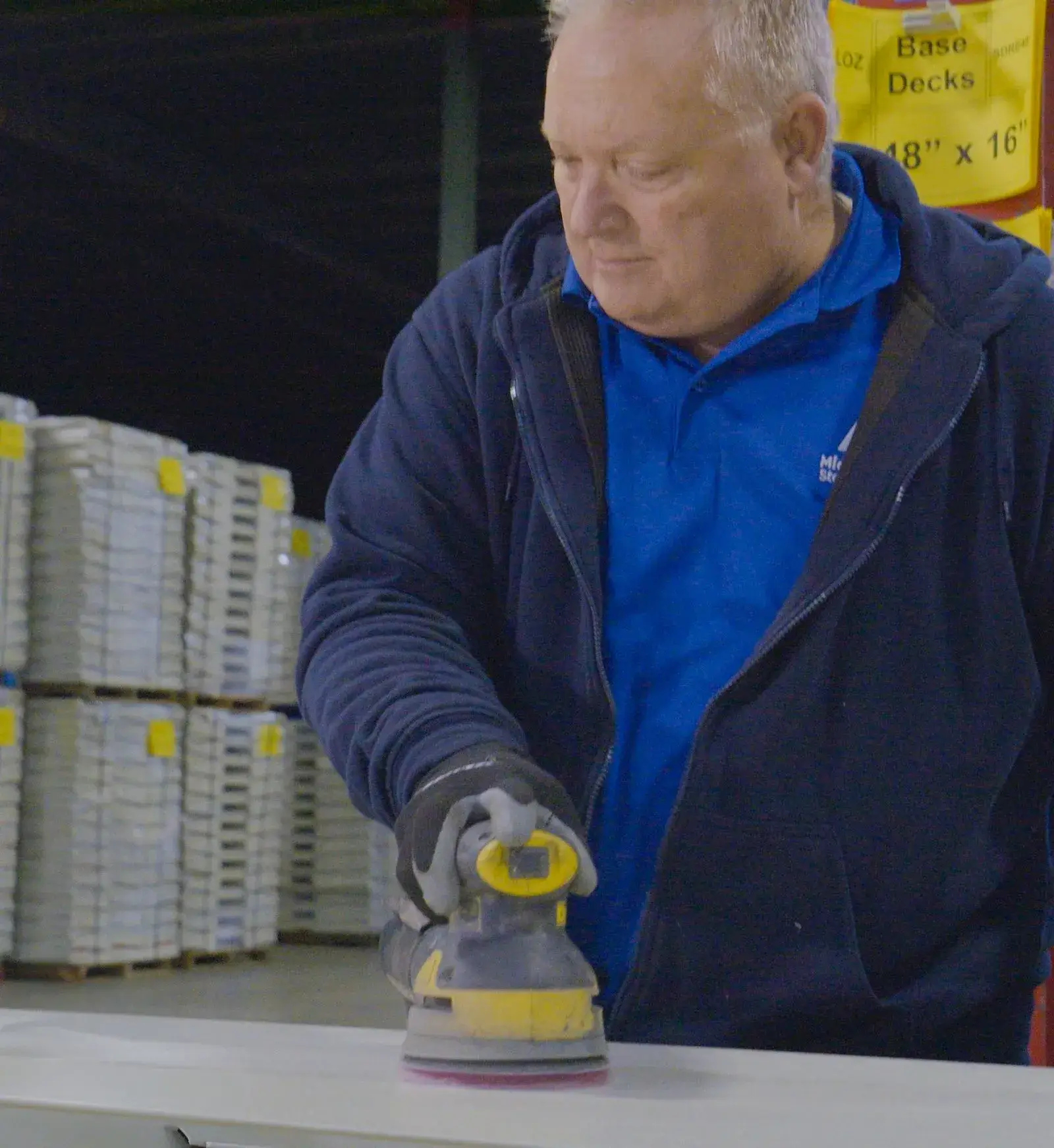 A person sanding a surface in a warehouse environment with stacked shelving in the background.