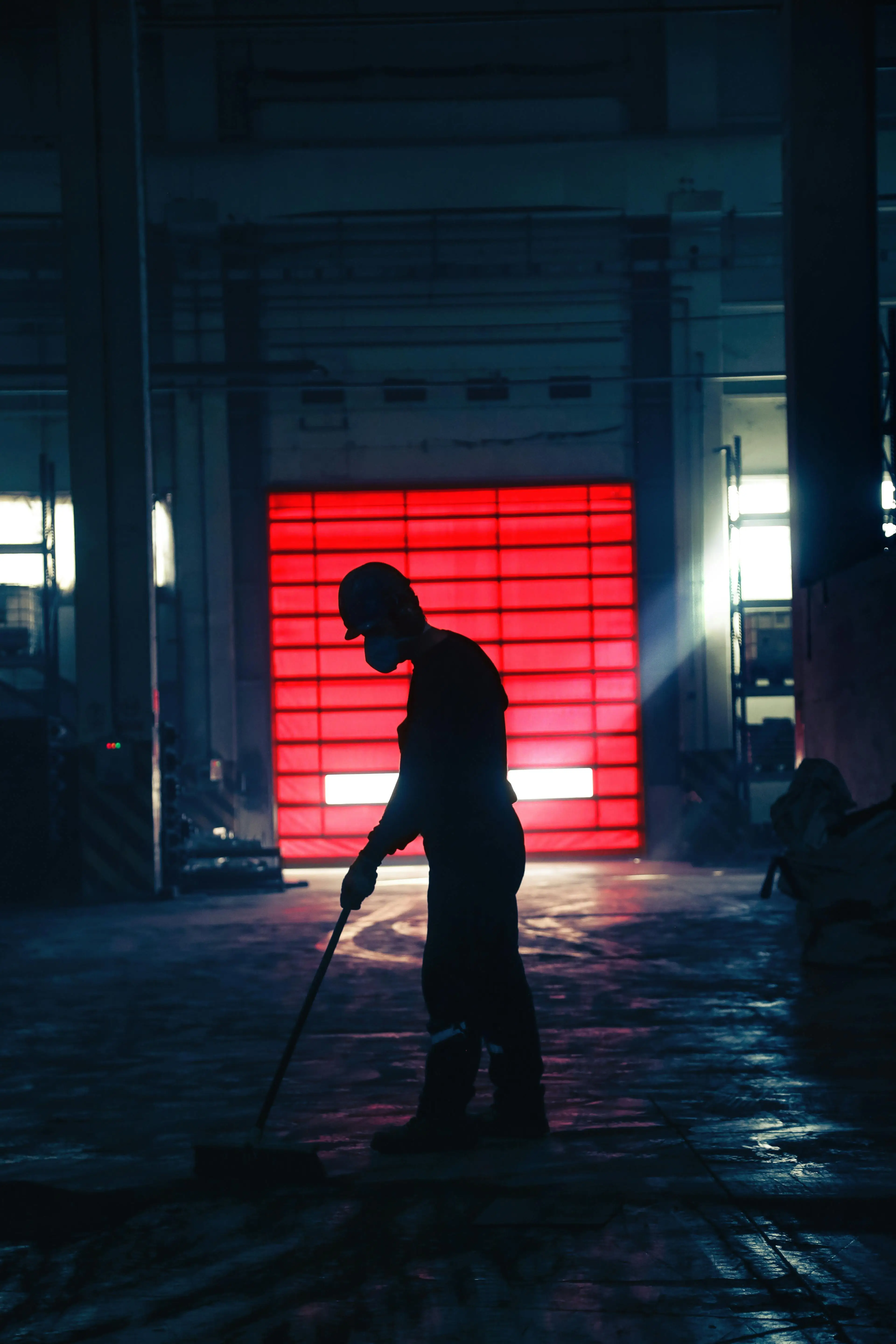 Silhouette of a person sweeping a warehouse floor in front of a large red door, with ambient lighting.