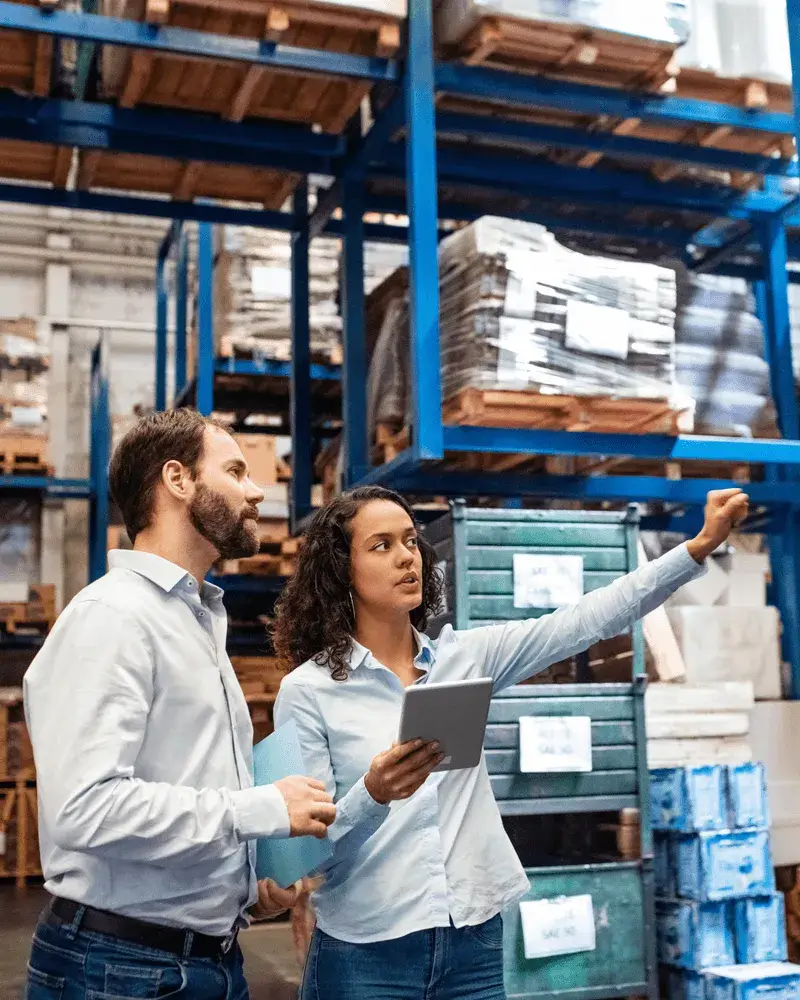 Two employees having a discussion in a warehouse with pallet racks and inventory in the background.