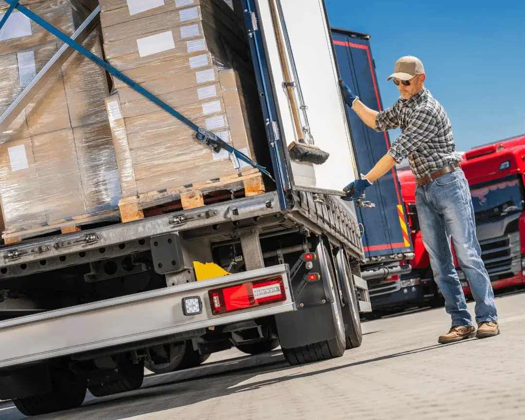 A worker wearing a cap and gloves is closing the back of a truck loaded with palletized boxes. The scene is outdoors with other trucks visible in the background.