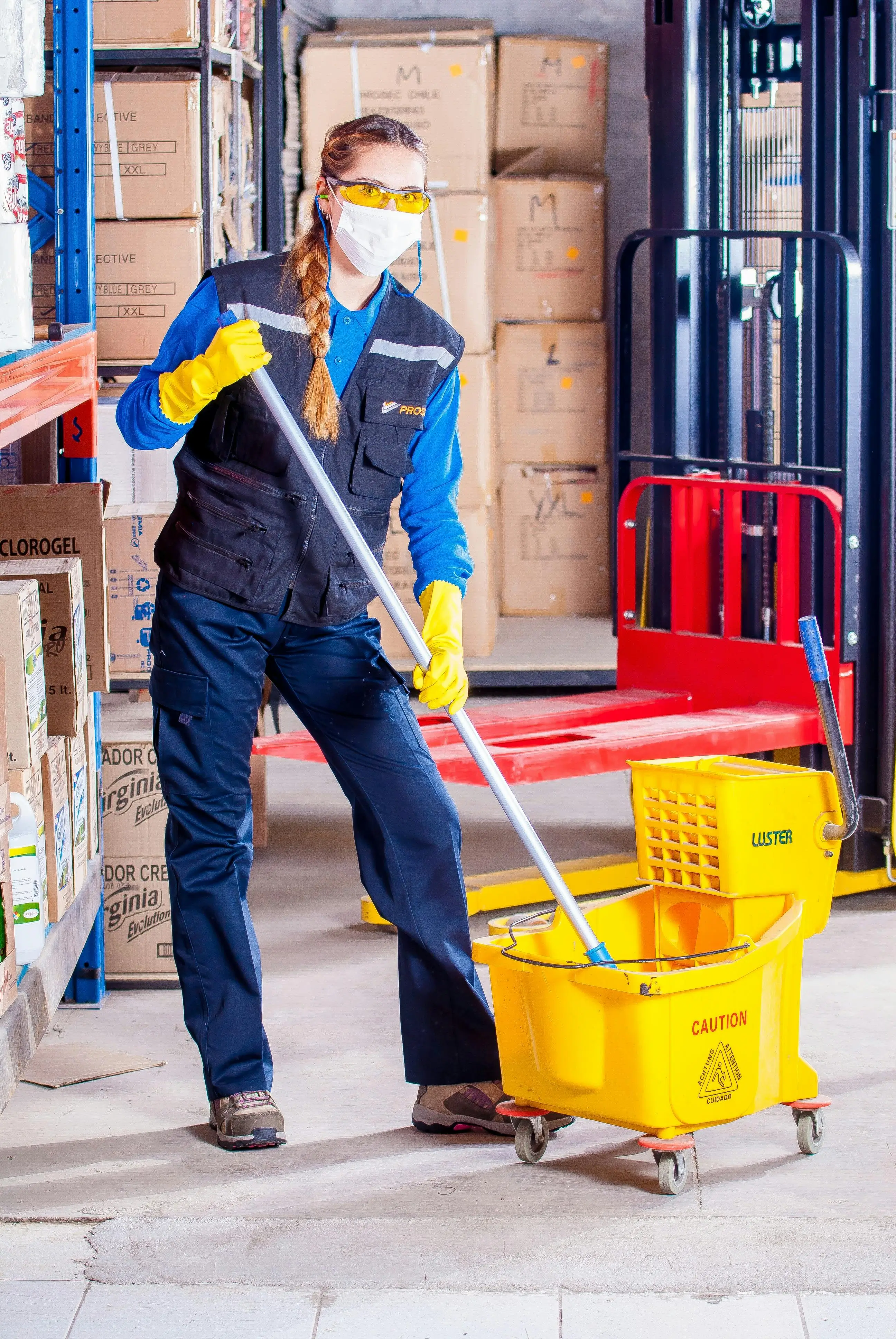 Person wearing safety gear mopping a warehouse floor with boxes and shelves in the background.