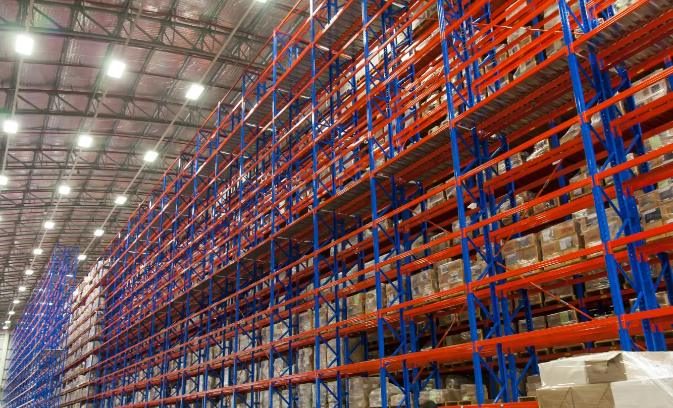 Large warehouse with tall blue and red pallet racking filled with boxed goods under bright lighting.