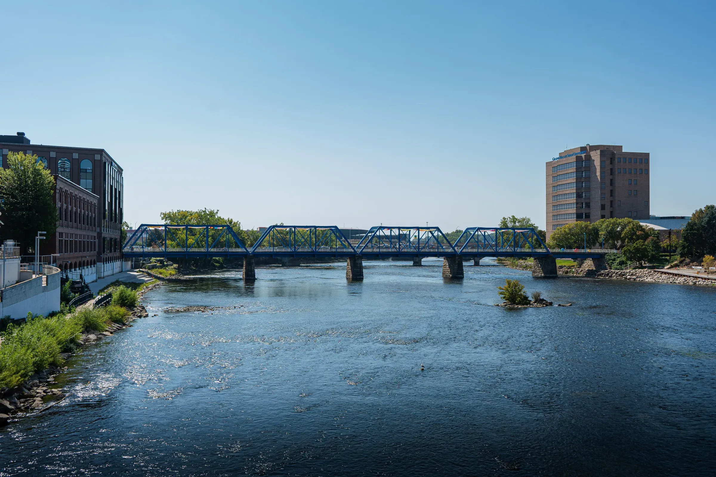 Blue steel truss bridge spanning a calm river with grassy banks, brick buildings on either side, and trees lining the waterway under a clear sky